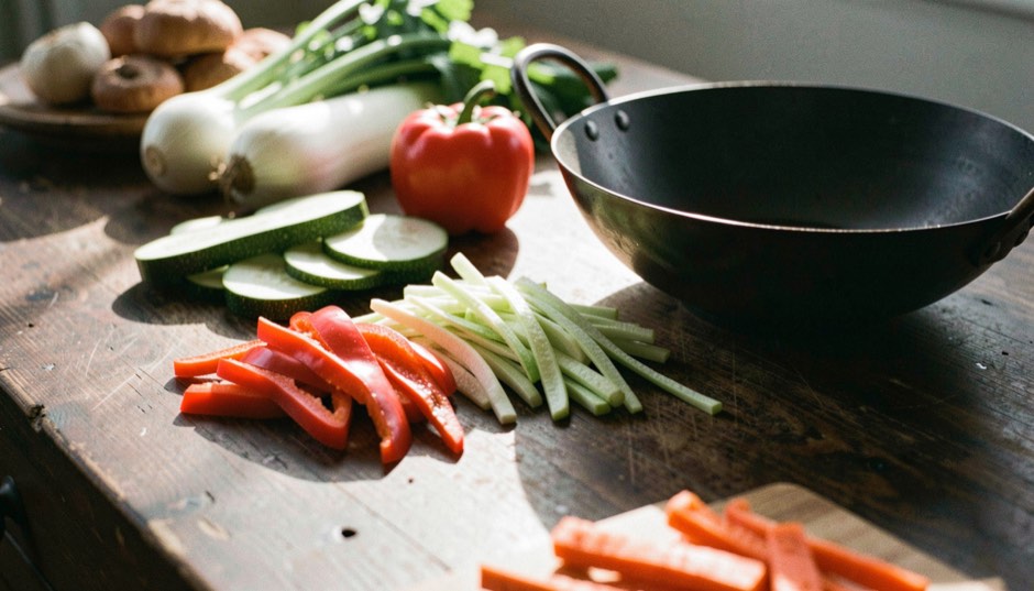 Un plan de travail en bois sombre couvert de légumes taillés, rondelles de courgette, lanières de poivron rouge, bâtonnets de carotte, poireaux et champignons, à côté d’un wok en acier noirci, lumière rasante de fin de journée